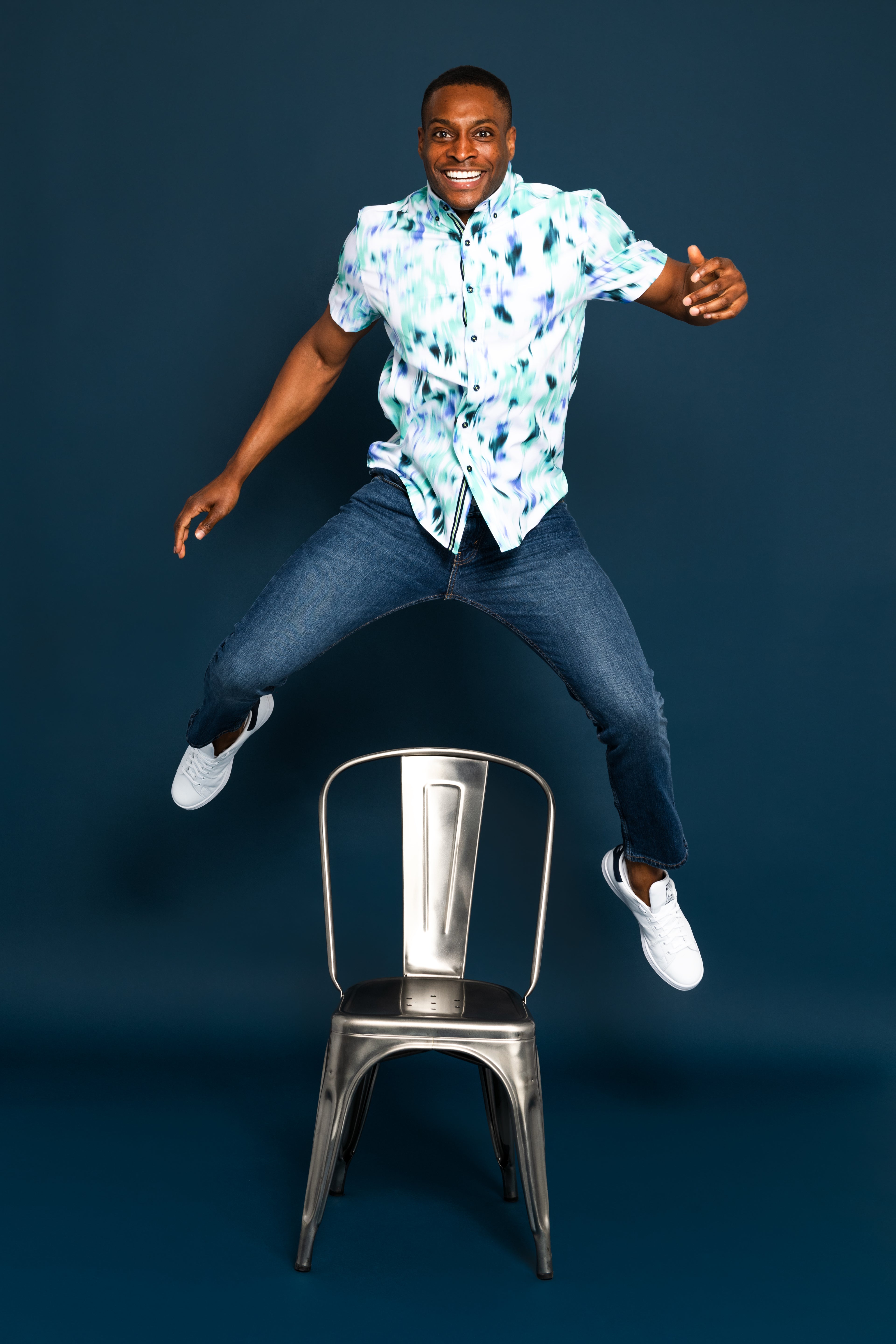Man in a colorful shirt and jeans sitting on a metal chair against a dark blue background