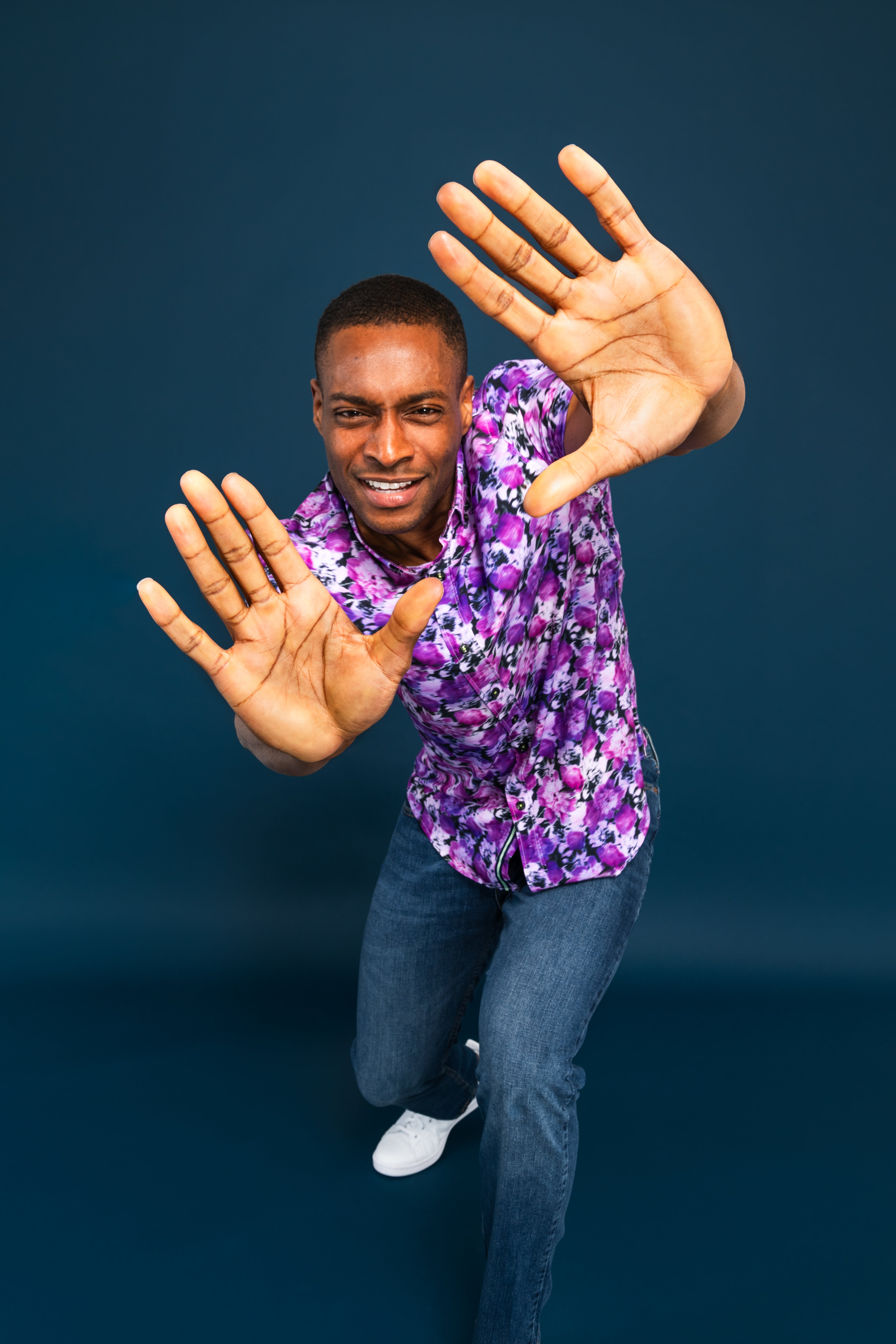 Man in a purple floral shirt and blue jeans standing against a dark blue background with his hands outstretched.