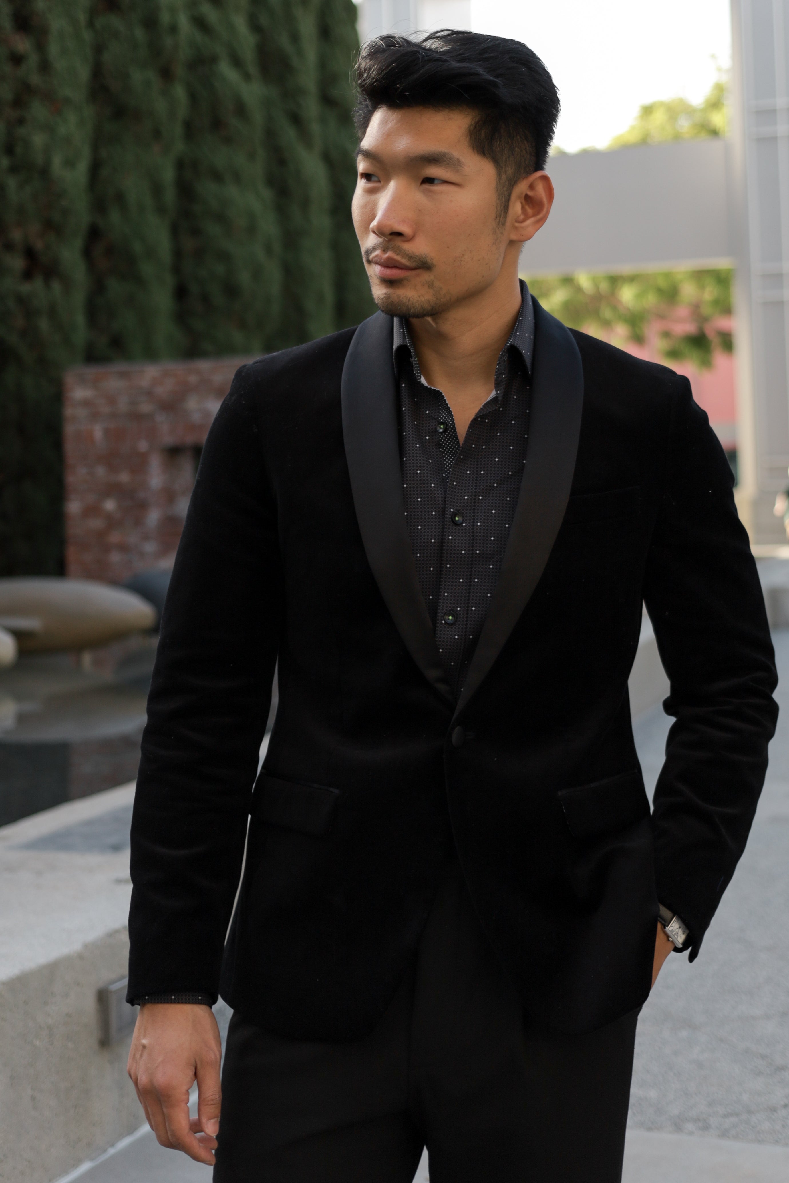 Man wearing a black tuxedo with a patterned shirt outdoors.