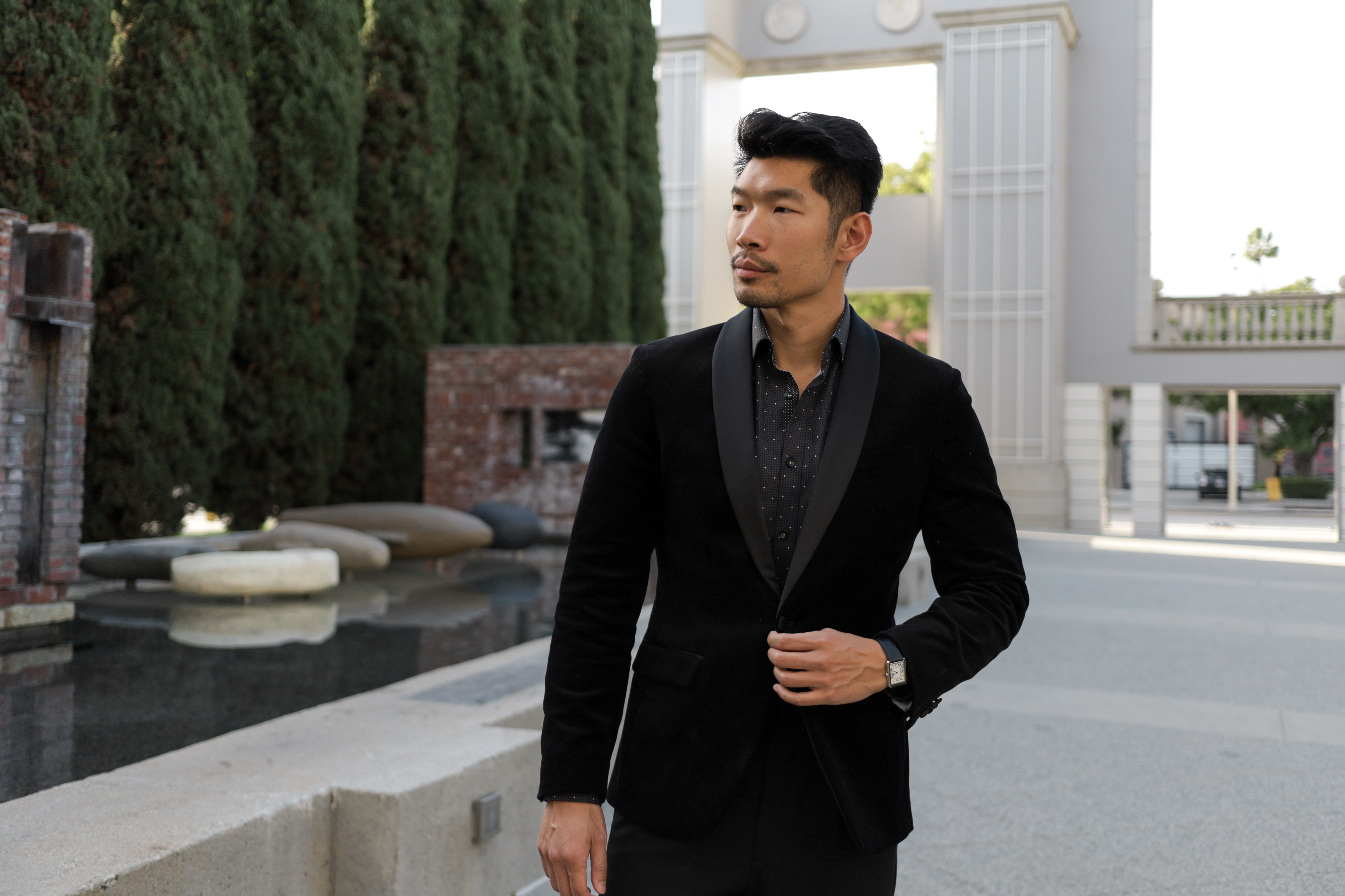 Man wearing a black tuxedo standing outdoors near a fountain.